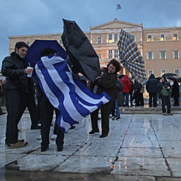 Fitch declassa la Grecia a C. Nella foto alcuni manifestanti, con una bandiera greca, cercano di tenere gli ombrelli sotto la pioggia e le raffiche di vento, di fronte al parlamento greco durante una manifestazione di protesta ad Atene (AP Photo)