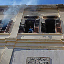 Incendio all'Istituto d'Egitto, corsa contro il tempo per salvare preziosissimi testi antichi. Nella foto un giovane manifestante egiziano lancia pietre da una finestra del complesso scientifico durante gli scontri con i soldati dell'esercito vicino a piazza Tahrir al Cairo (AP Photo)